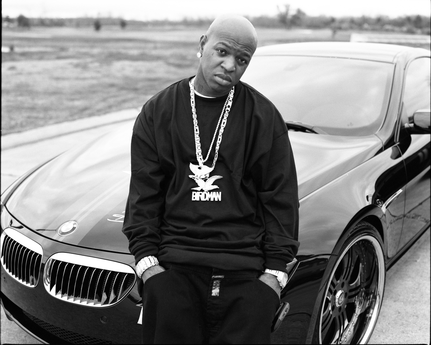 Black and white photograph of Birdman standing in front of a car and wearing a large metal chain