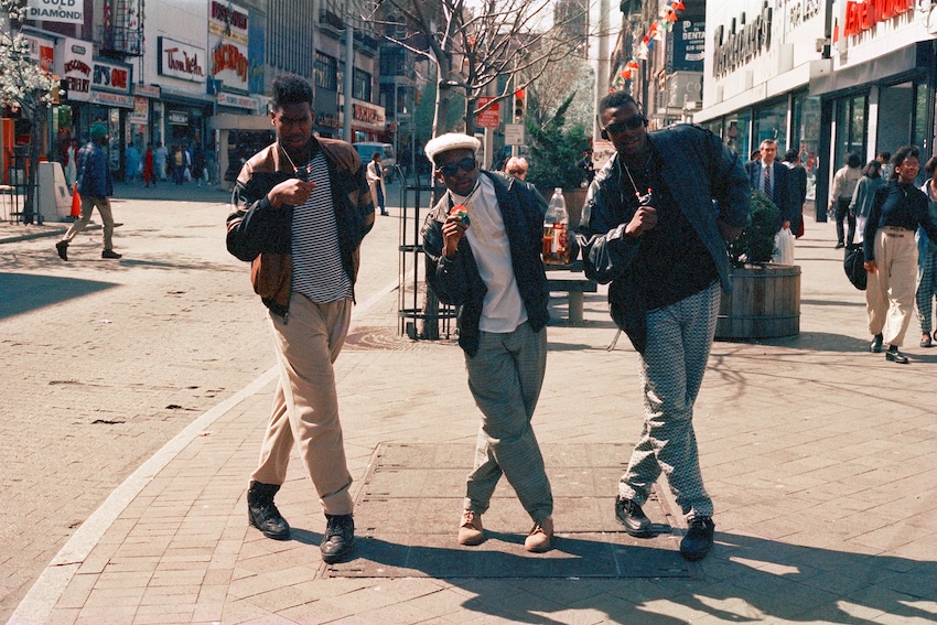Photograph of three men standing with their legs crossed, facing the camera on a street Brooklyn, NYC.