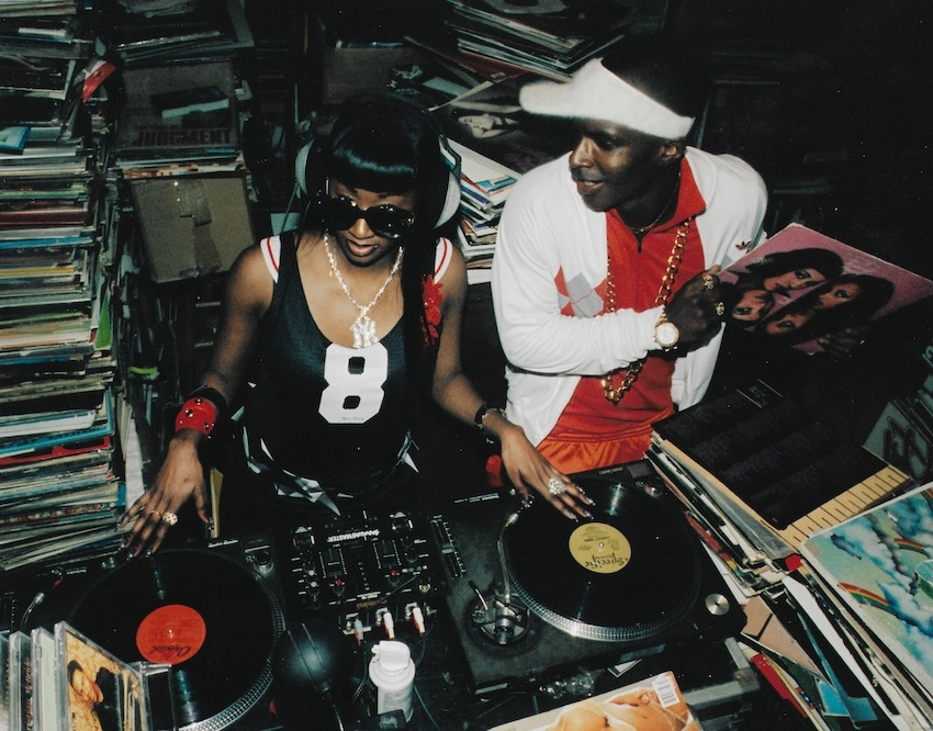 Photograph of two djs standing above decks, with lots of records behind them in the background.