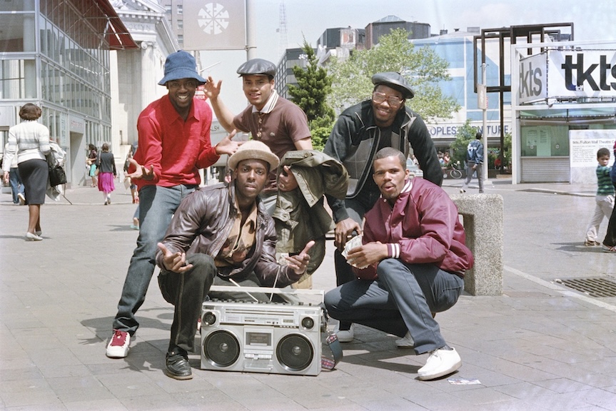 Photograph of a group of five men standing above a 90s style boombox on a street